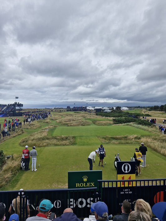 A player and caddies walk a links fairway at The Open Championship, Rolex leaderboard visible in the background, grey skies overhead and a large gallery of spectators lining the course