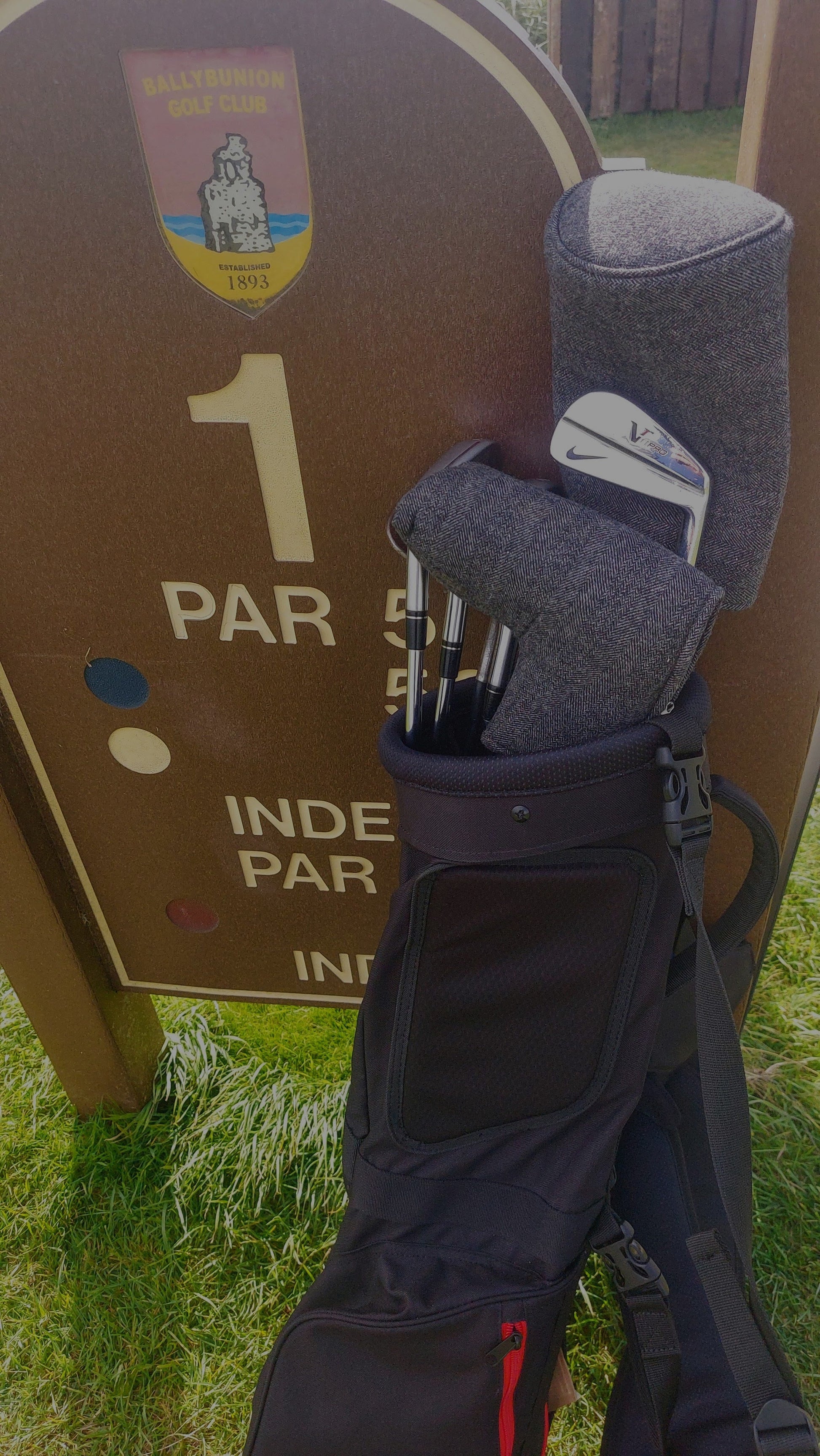 A set of golf clubs leaning up against a Ballybunion Golf Club sign. The clubs have charcoal barrel headcovers.