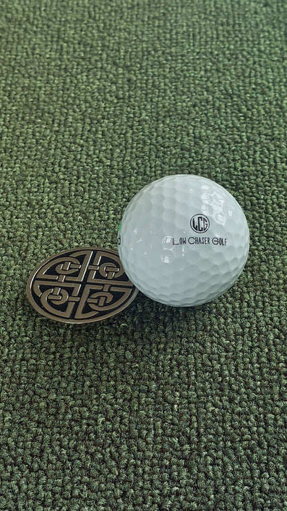 Golf ball marker, with a celtic shield knot design, marking a Low Chaser Golf ball. Both sitting on artificial grass